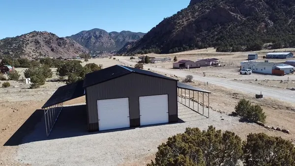 Brown steel building with two white roll-up doors and lean-tos on both sides below rocky hills.