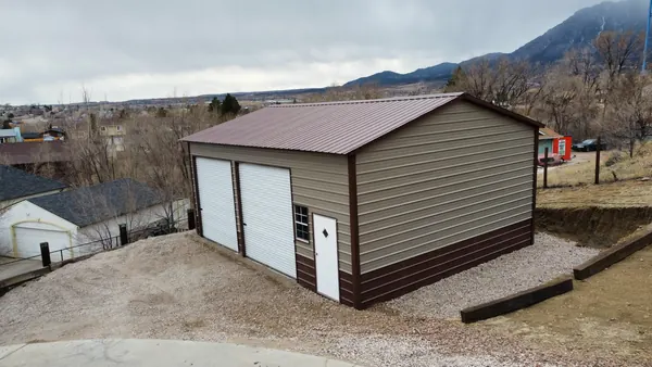 Detached tan steel garage with a brown roof and two roll-up doors on a sloped residential lot.