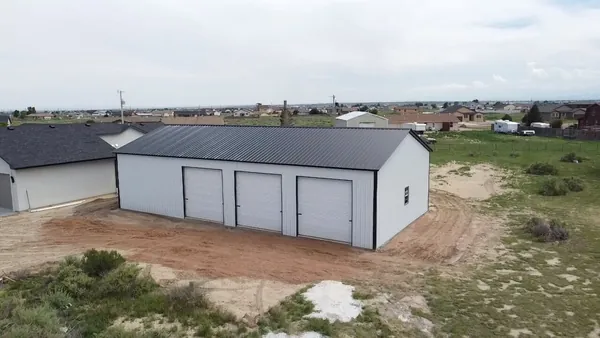 Gray steel garage with a black roof and three white roll-up doors beside a house, aerial view.