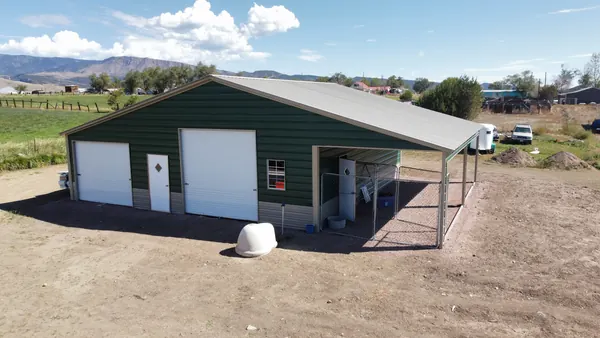 Green steel building with roll-up doors and a covered side lean-to on a rural lot, mountains on the horizon.