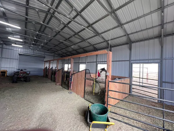 Inside a steel horse barn with a roll-up door, open center aisle and horse stalls along one wall.