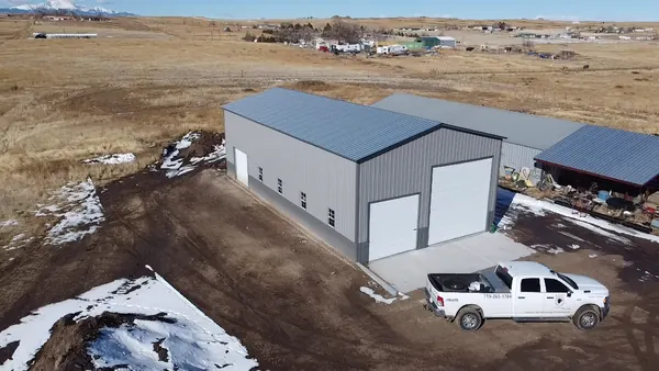 Large gray steel workshop with two roll-up doors and a white pickup truck, patches of snow on the ground.