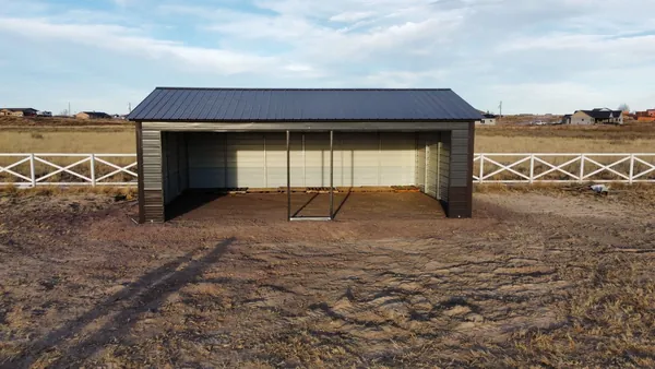 Front view of an open-front steel loafing shed with a dark roof and brown wainscot in a fenced pasture.