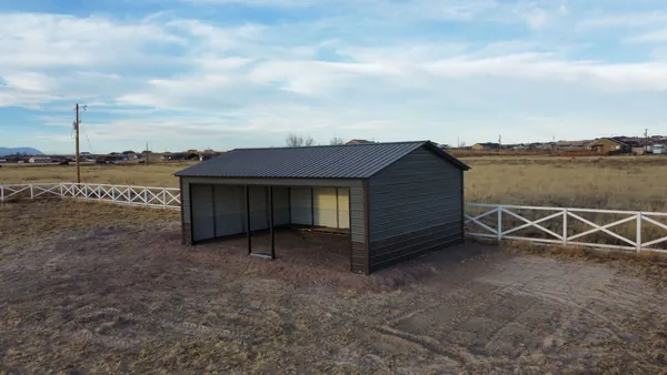 Steel loafing shed with one open side and a dark roof inside a white crossbuck-fenced pasture.