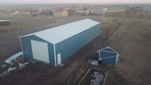 Long blue-gray steel building with a white roll-up door on a fenced rural lot, light snow on the ground.