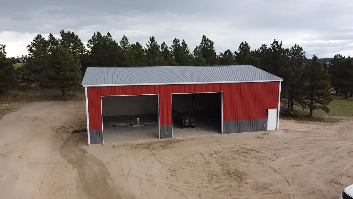 Red steel workshop with gray wainscot and two open bays, set in a clearing among pine trees.