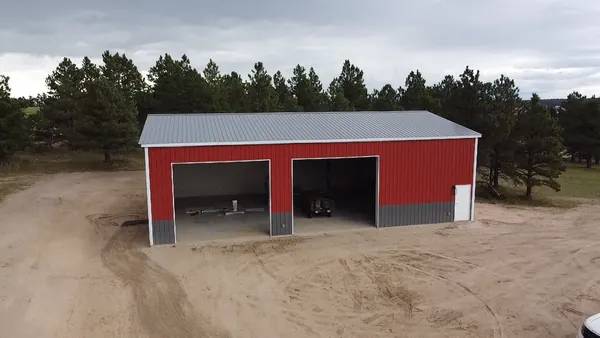 Red steel workshop with gray wainscot and two open bays, set in a clearing among pine trees.