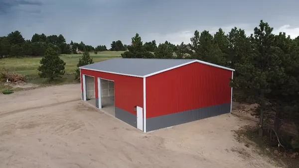 Red steel workshop with two roll-up doors and gray wainscot in a clearing surrounded by pines.