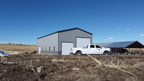 Gray steel building with roll-up doors and a side lean-to, white pickup truck parked in front.