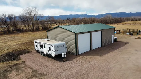 Tan steel RV garage with a green roof and two roll-up doors, travel trailer parked beside it, foothills behind.