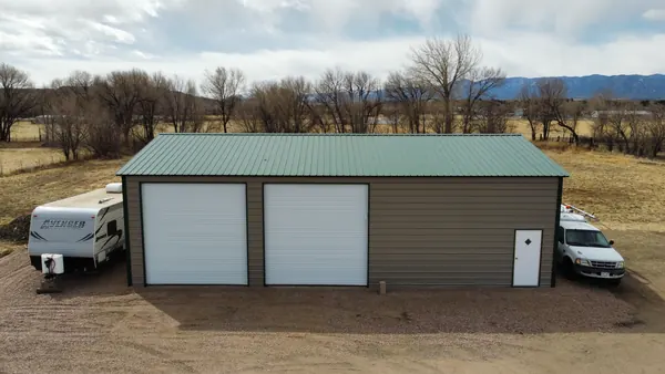 Tan steel garage with a green roof and two tall roll-up doors, a travel trailer and truck parked alongside.