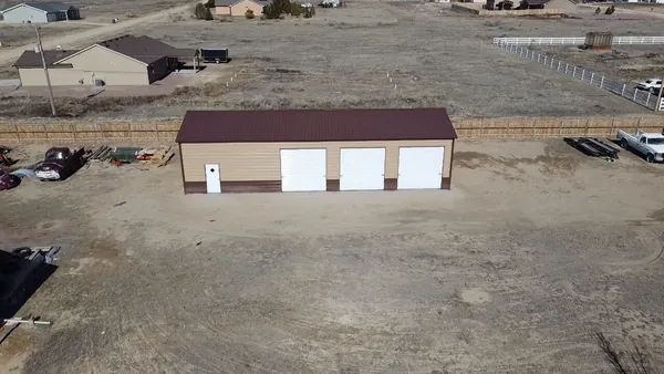 Tan steel garage with a brown roof, three white roll-up doors and a walk-in door, aerial view.