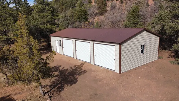 Tan steel garage with three white roll-up doors and a brown roof among pine trees, aerial view.