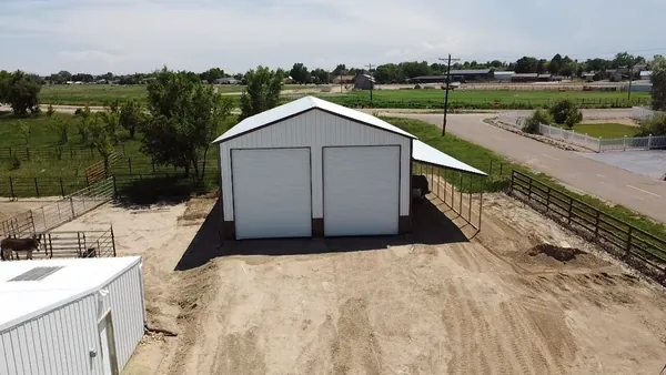 White steel garage with two roll-up doors and a covered side lean-to next to a corral, aerial view.