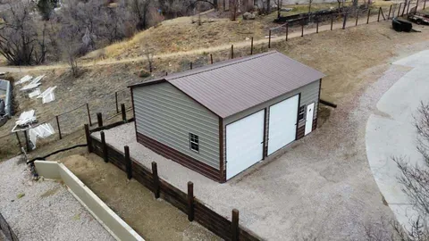 Tan steel garage with a brown roof and two white roll-up doors on a fenced, sloped lot