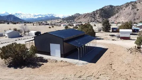 Black steel garage with white roll-up doors and a covered lean-to, rocky hills behind