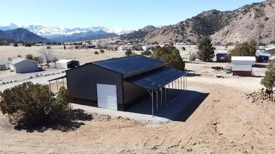 Black steel garage with white roll-up doors and a covered lean-to, rocky hills behind