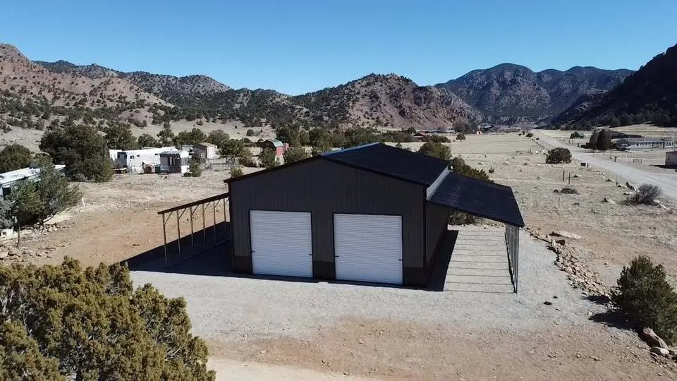 Black steel shop with two roll-up doors and lean-tos on a gravel pad below the mountains