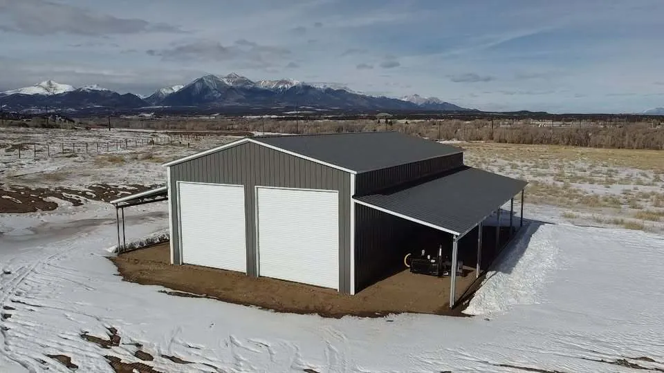 Gray steel building with two roll-up doors and full-length lean-tos in snow, peaks behind