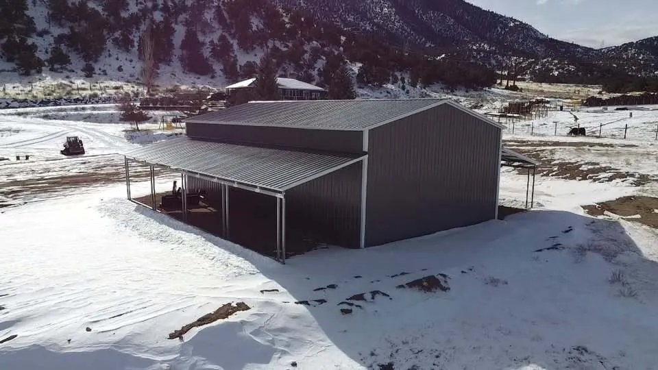 Side view of a gray steel building with a full-length lean-to on a snowy hillside lot