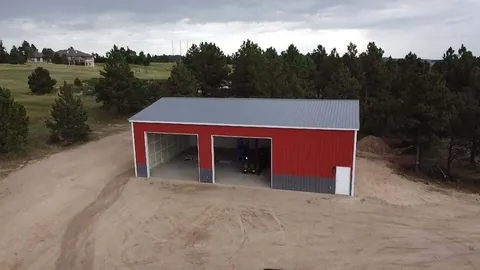 Red steel garage and shop with gray wainscot and two open bays, set among pine trees